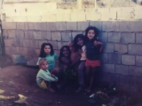 Foto antiga de um grupo de seis crianças sentadas e em pé em frente a um muro de blocos de concreto.