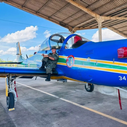 Uma militar fardada sentada na asa de um avião azul da Esquadrilha da Fumaça, dentro de um hangar.