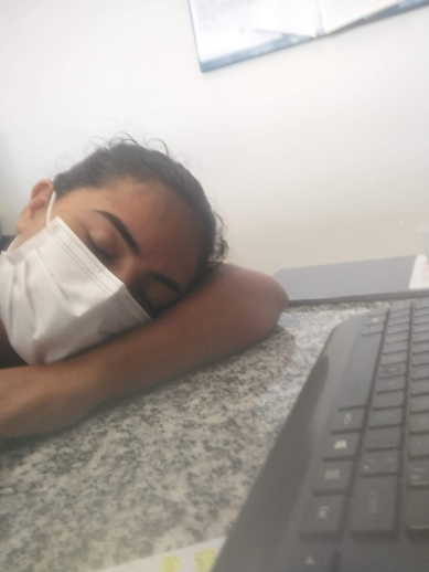 Mulher usando máscara branca de proteção descansa a cabeça sobre o braço em uma mesa ao lado de um teclado.