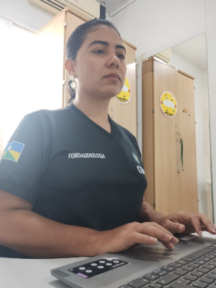 Mulher de cabelos presos e camiseta preta com a inscrição "Fonoaudiologia" sentada à frente de um notebook em um escritório.
