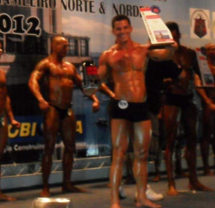 Fisiculturista sorridente posa em um palco segurando um troféu.
