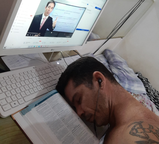 Homem dormindo sobre um livro aberto em frente ao computador, que exibe uma videoaula