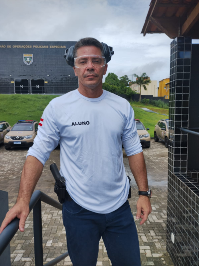 Homem em treinamento policial, usando equipamentos de proteção, posicionado em frente a um batalhão com viaturas.