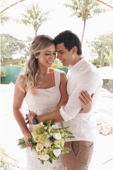 Casal de noivos se abraçando e sorrindo, com as testas encostadas. A noiva segura um buquê de flores