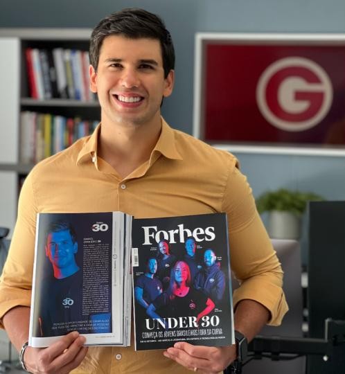 Homem sorridente segurando exemplares abertos da revista Forbes Under 30