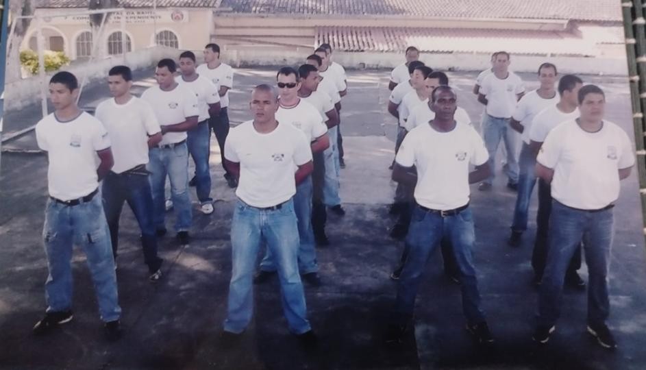 Grupo de homens em formação militar, vestindo camisetas brancas e calças jeans, parados em posição de descanso em um pátio