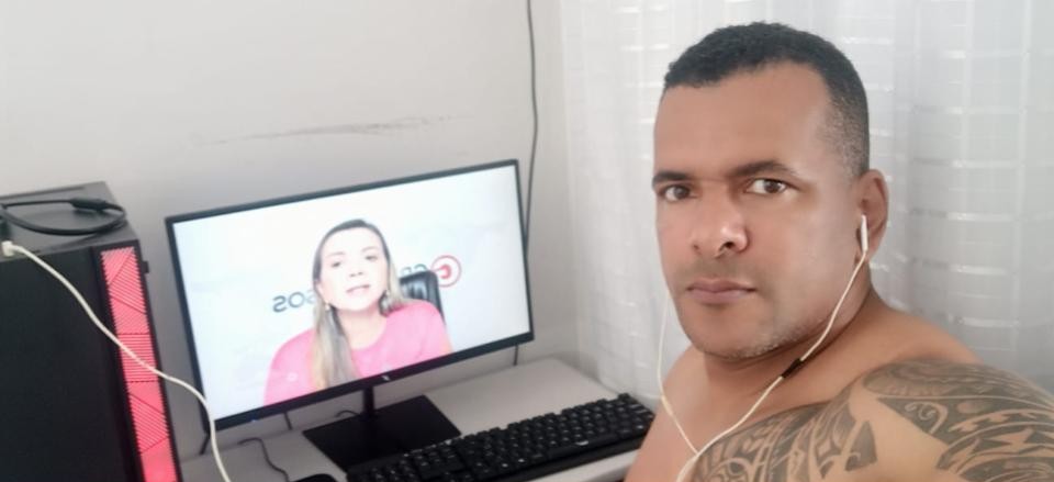 Homem com fones de ouvido posicionado ao lado de um computador, onde o monitor exibe uma videoaula