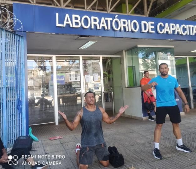Homem de regata ajoelhado no chão com os braços abertos e um homem de camisa azul em pé ao seu lado, em frente à fachada de um laboratório de capacitação.