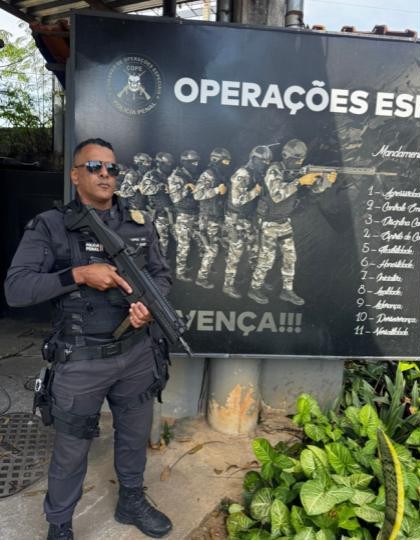 Policial fardado e armado posicionado à frente de um painel de "Operações Especiais"