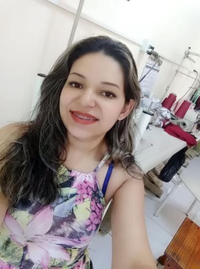 Mulher tira selfie sorridente e ao fundo estão linhas e máquina de costura. 