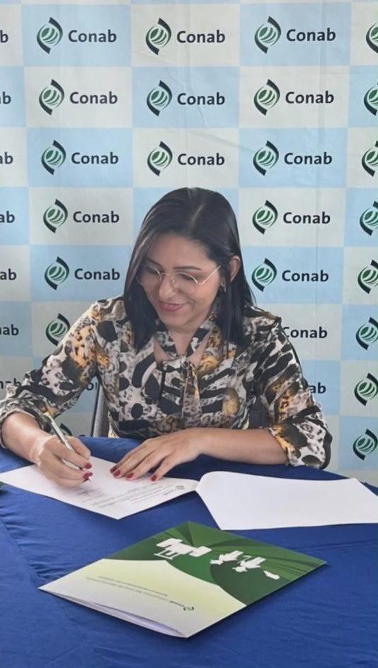 Mulher sorridente assinando documentos. Ao fundo, um painel escrito Conab